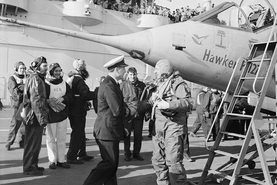 Mr. A. W. Bedford, chief test pilot of Hawker Aircraft Ltd, is welcomed aboard HMS ARK ROYAL by Commander A. R. Rawbone, after he had made the first ever vertical landing on an aircraft carrier at sea by a jet aircraft, the prototype of the Harrier, P.1127. (IWM A 34712)