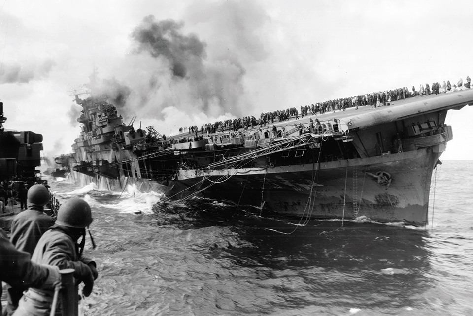 Listing heavily but still afloat after taking bomb hits on March 19, 1945, USS Franklin burns as its crew gathers on the flight deck. (National Archives)