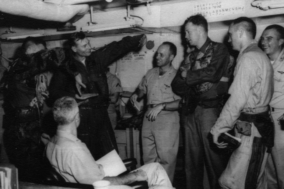 Lt. j.g. Charlie Hartman, in camoflage, and mission leader Lt. Cmdr. Edwin Greathouse describe to Rear Admiral William Bringle, seated, the downing of a MiG on June 20, 1965. Second from the right is Lt. Clinton Johnson. (National Archives)