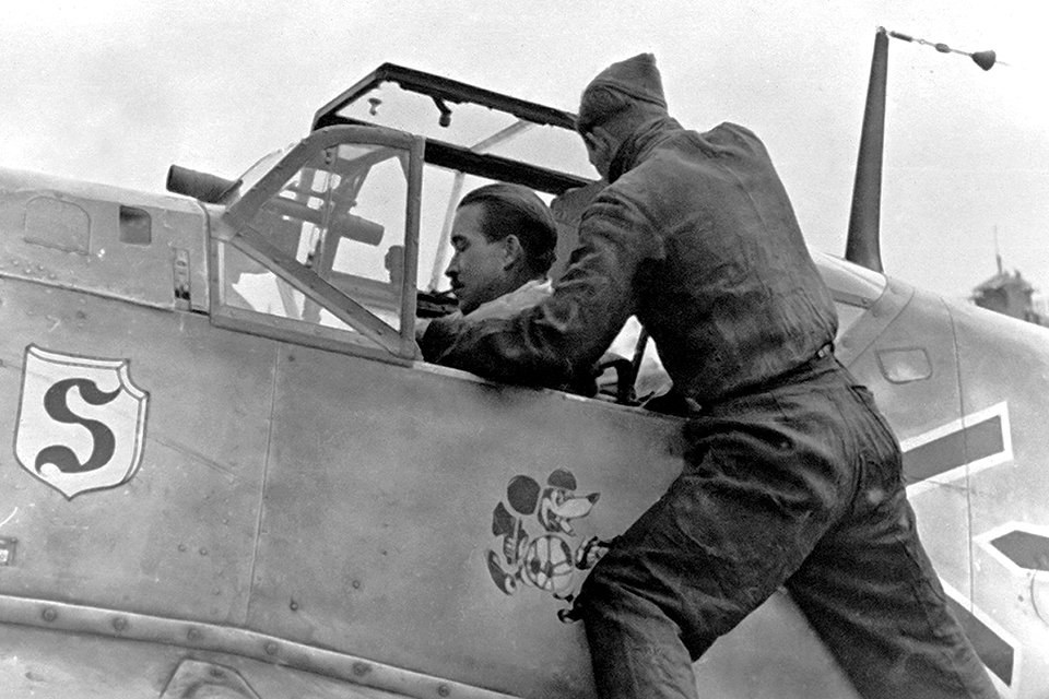 Galland prepares for a mission in his Me-109E, which sports a telescopic gunsight and his signature emblem of a weapon-toting, cigar-smoking Mickey Mouse. (Courtesy of Wolfgang Muehlbauer)