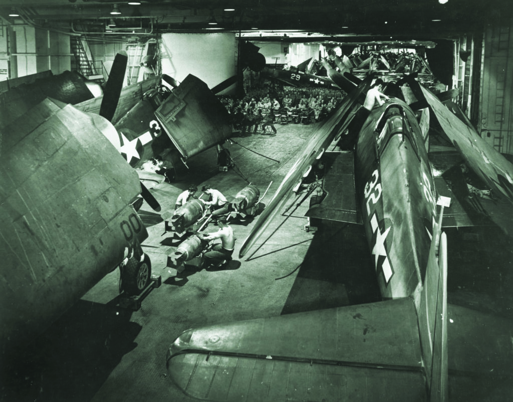 Hellcat fighters surround a group of ordnancemen preparing bombs on Yorktown’s hangar deck in late 1943. Behind them, crewmen assemble to watch a film. (Charles E. Kerlee/U.S. Navy)
