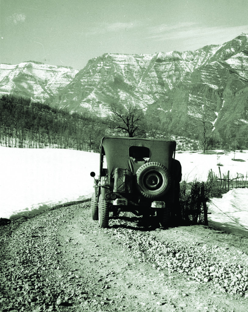 An army jeep pauses before the snow-dusted expanse of Italy’s Riva Ridge (above). Below: troops of the 87th Regiment’s F Company—one holding a German helmet—assemble before a bombed building. (The Denver Public Library, TMD-374) 