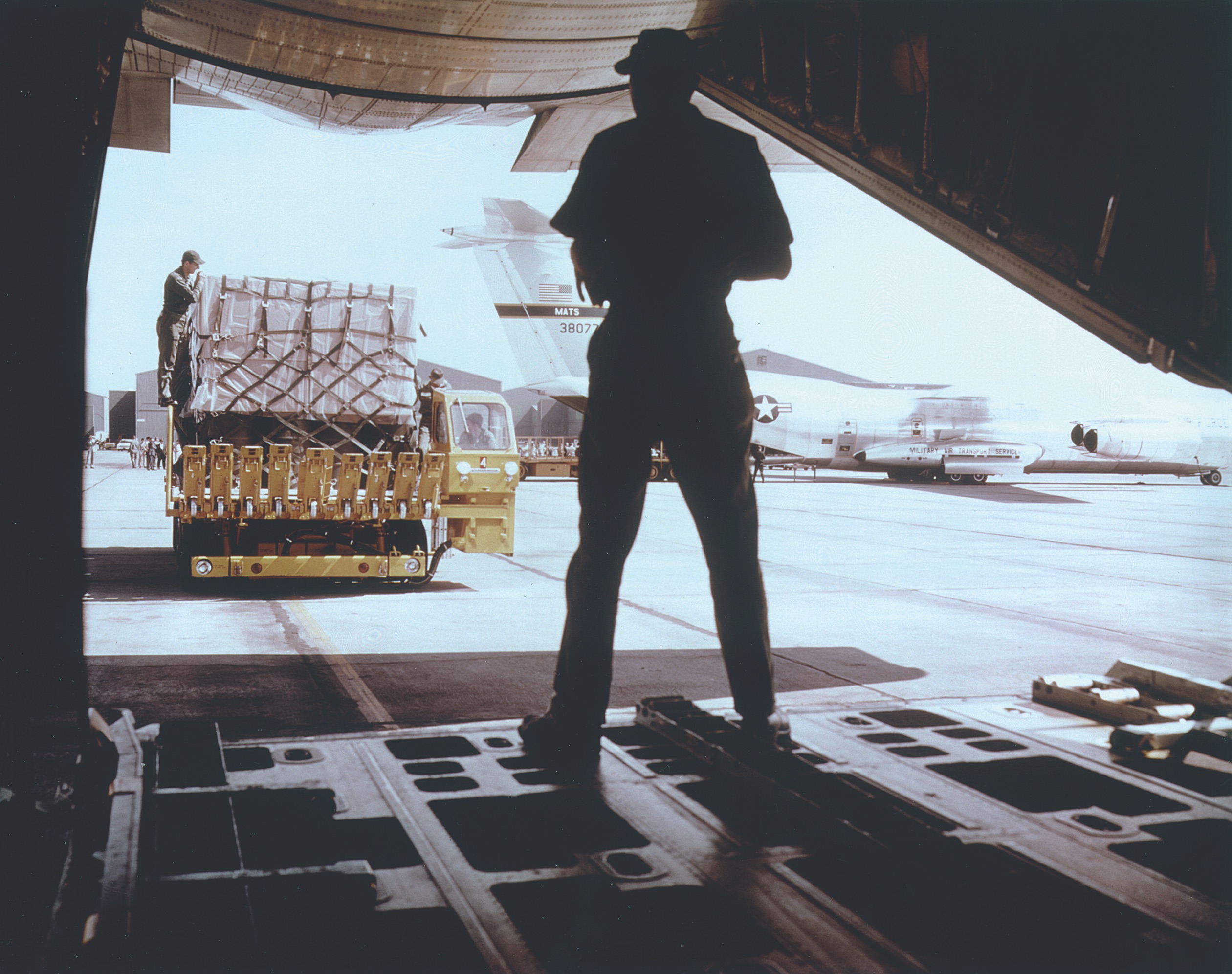 A loadmaster in the rear of a C-141 directs the driver of a cargo-loading vehicle as it moves toward the plane’s lowered ramp. / U.S. Air Force