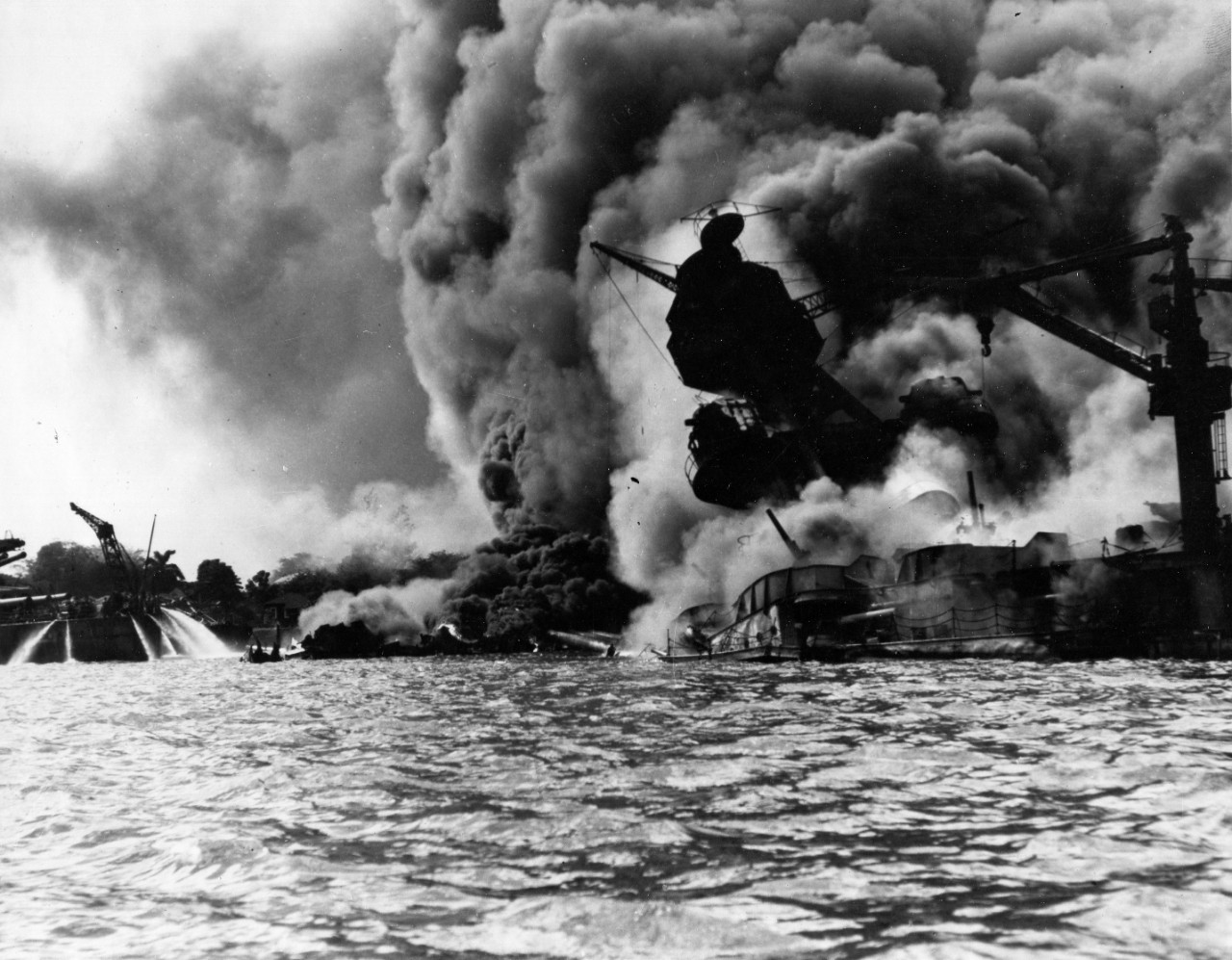 USS Arizona sinking and burning furiously on December 7, 1941. Her forward magazines exploded when she was hit by a Japanese bomb. At left, men on the stern of USS Tennessee are using fire hoses on the water to force burning oil away from their ship. (National Archives)