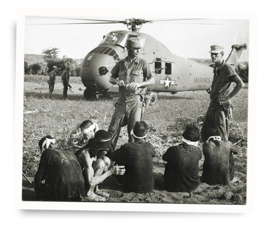 Photo of Viet Cong prisoners await transport near a UH-34D Seahorse helicopter with Marines standing guard during Operation Starlite in August 1965.
