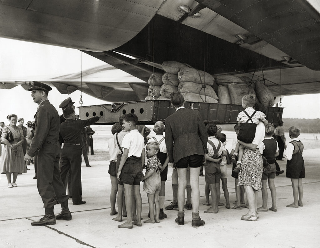 Photo of A group of refugees watches in anticipation as a platform teaming with flour sacks descends from the cargo hold of the mammoth Douglas C-47 Globemaster, the largest cargo aircraft of its kind during the time of the Berlin Airlift.