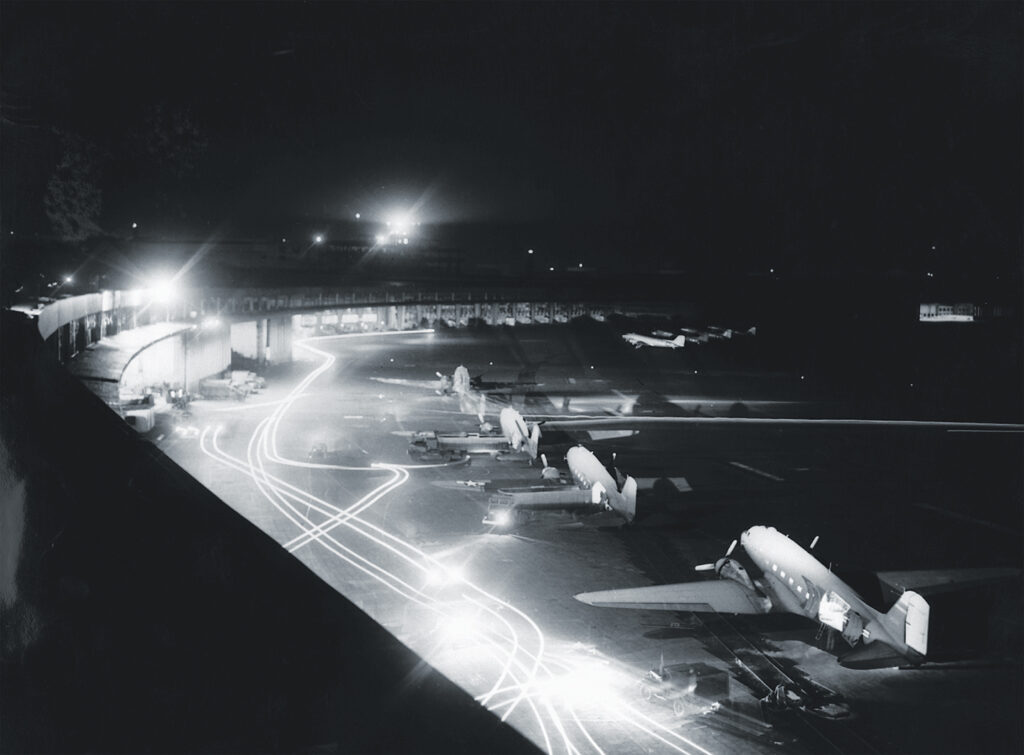 Photo of A fleet of Douglas C-47 Skytrain cargo planes waits to deliver food supplies during the Berlin Airlift as trucks busily navigate the darkened tarmac.