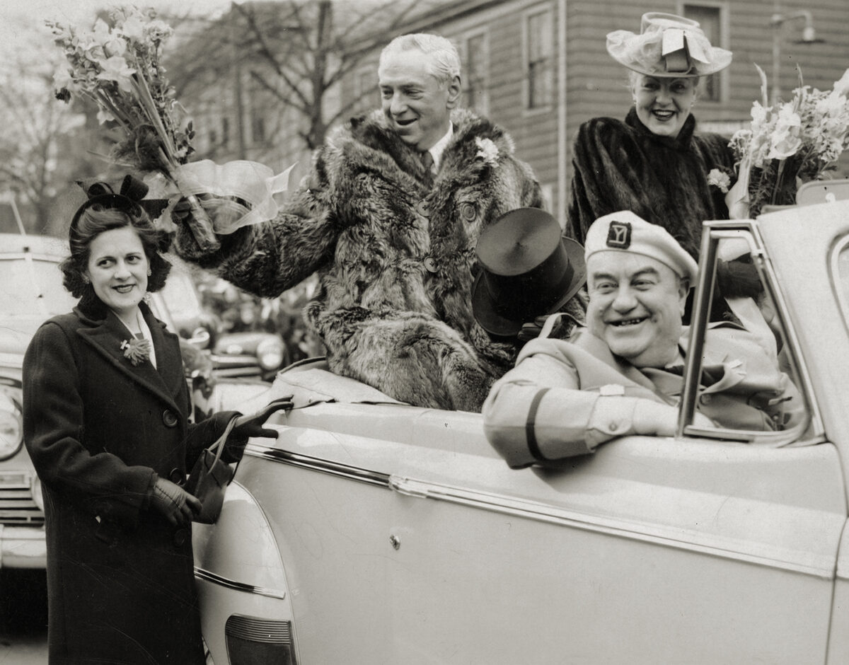 Photo of Mayor James Michael Curley, dressed in his raccoon coat, hands out flowers during South Boston's traditional Evacuation Day parade on March 17, 1947. Mayor Curley's wife, Gertrude, in a smart green hat with a pink ribbon is sitting to his left and Edward J. "Knocko" McCormack in his Yankee Division uniform is in front. The parade originally commemorated the day the British left Boston on March 17, 1776 and now it also honors St. Patrick, the patron saint of Ireland.