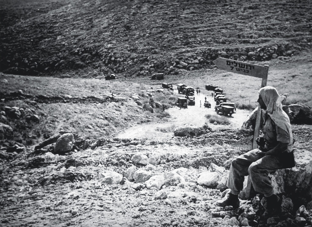 Photo of An armed Haganah soldier stands guard over the secret road which served as a lifeline to Jerusalem during the siege by thousands of Arab troops. A medical convoy proceeds cautiously along the road.