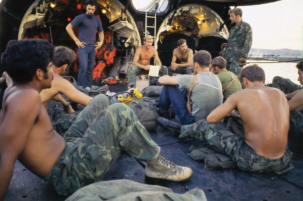 Photo of Lt Dry (in center holding paper and facing camera) briefs his SEAL Platoon “Alpha” on the deck of the submarine USS Grayback after his unit reported aboard in April 1972 at the U.S. Navy’s base at Subic Bay, Republic of the Philippines. Then-Chief Petty Officer Philip "Moki" Martin is visible at left in the foreground. Other members of the platoon included LT Robert W. Conger Jr., Samuel E. Birky, Timothy R. Reeves, Richard C. Hetzell, Eric A. Knudson, Robert M. Hooke, Frank Sayle, David Ray Hankins, John M. Davis, Michael J. Shortell, Barry S. Steele, and William B. Wheeler.
