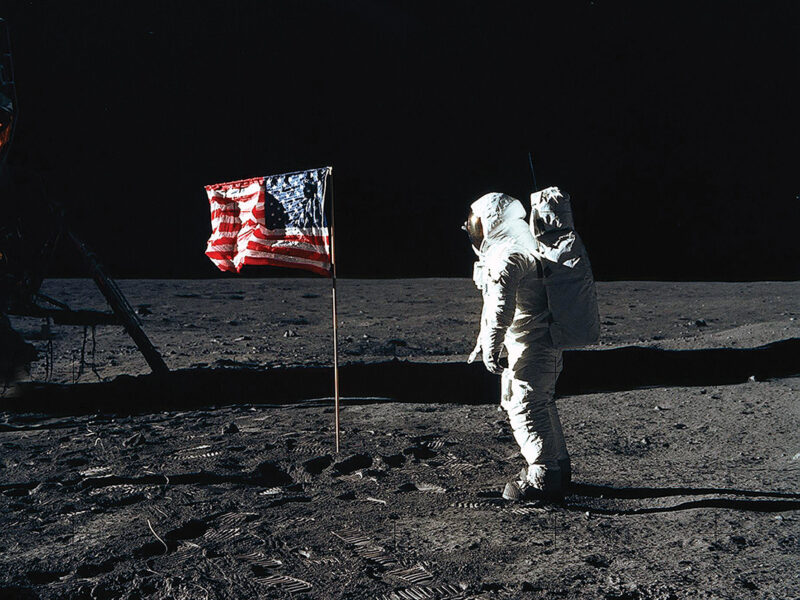 Photo of astronaut Edwin E. Aldrin Jr., lunar module pilot of the first lunar landing mission, poses for a photograph beside the deployed United States flag during an Apollo 11 extravehicular activity (EVA) on the lunar surface. The Lunar Module (LM) is on the left, and the footprints of the astronauts are clearly visible.