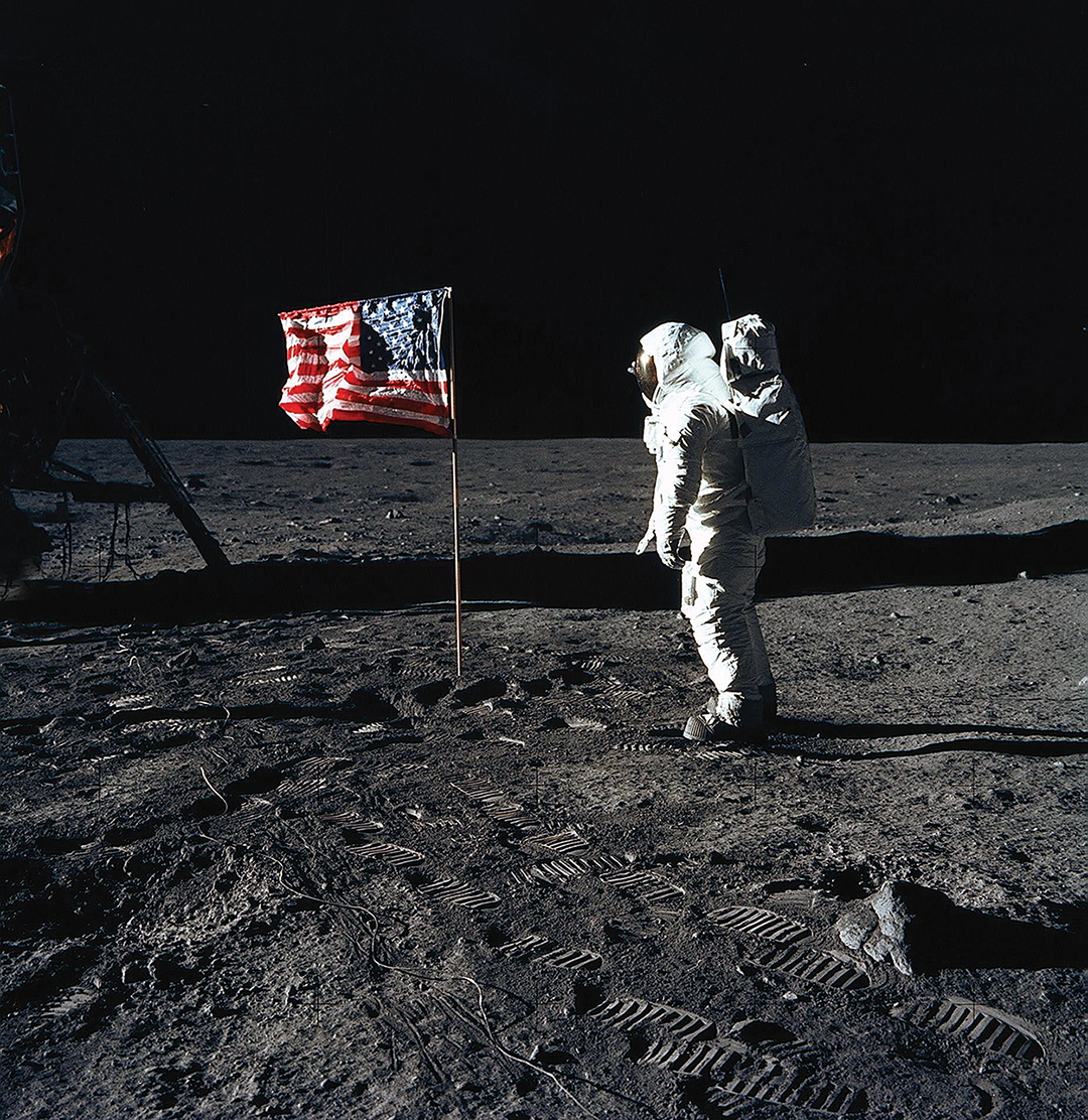 Photo of astronaut Edwin E. Aldrin Jr., lunar module pilot of the first lunar landing mission, poses for a photograph beside the deployed United States flag during an Apollo 11 extravehicular activity (EVA) on the lunar surface. The Lunar Module (LM) is on the left, and the footprints of the astronauts are clearly visible.